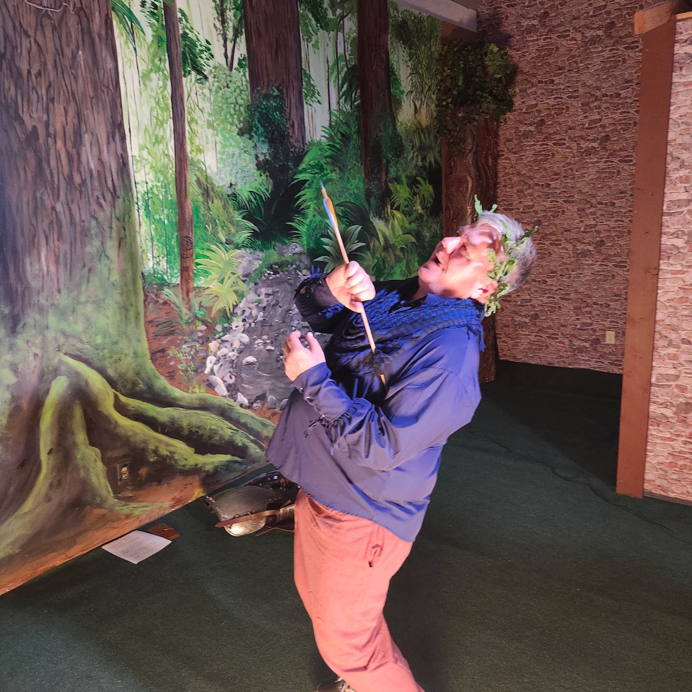 Toby Leonard in costume with a leafy crown, hamming it up with a prop arrow in the forest-themed room at Kato Escape in Mankato