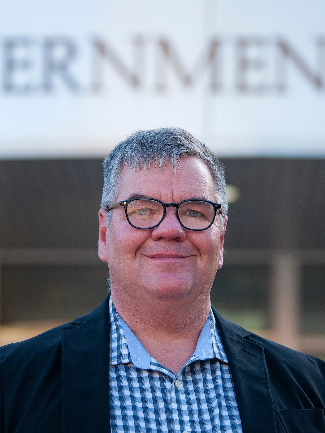 Toby Leonard in a dark blazer and checked shirt, smiling outside the Government Center