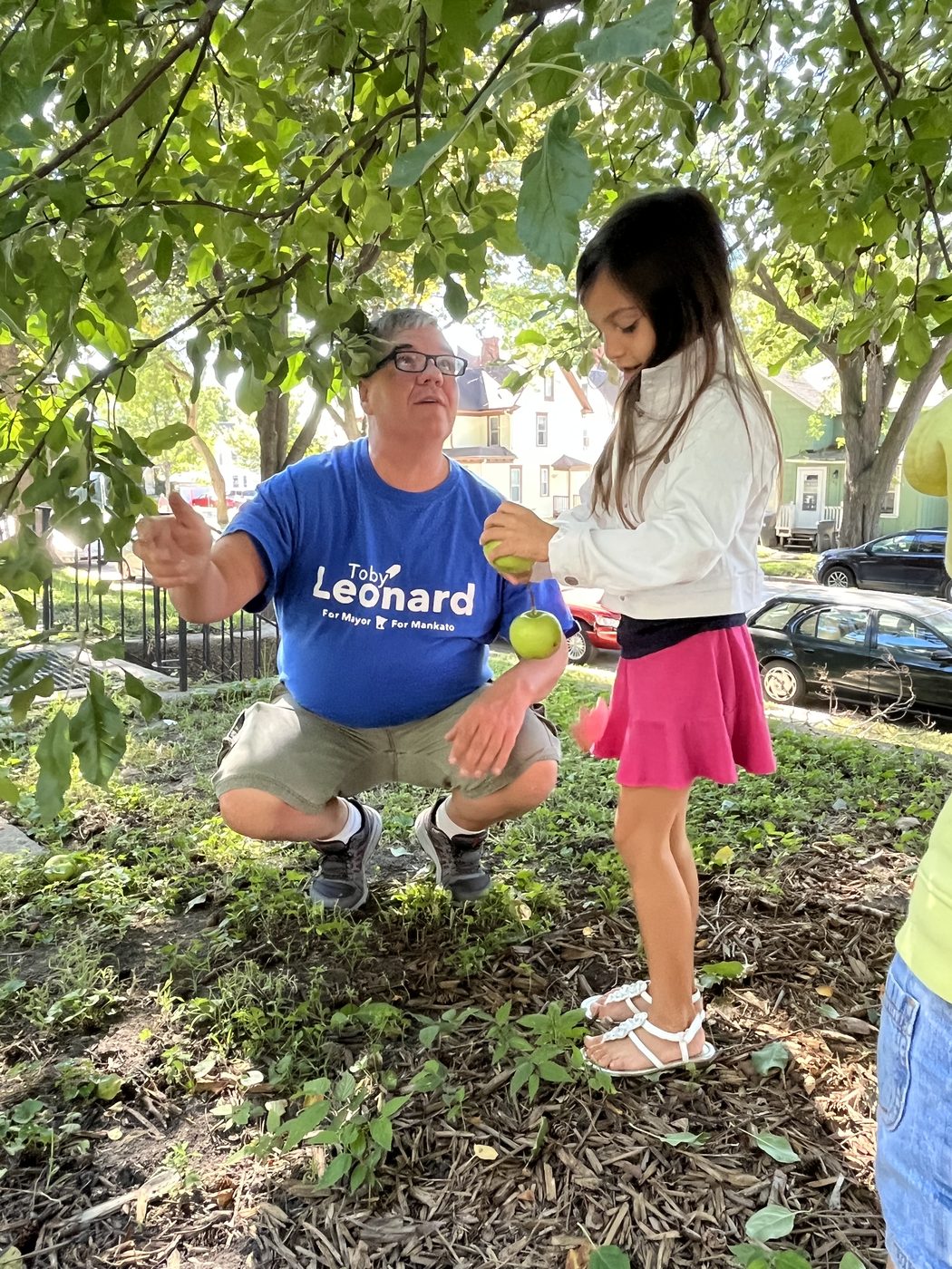 Toby Leonard crouching under an apple tree, handing a green apple to a young girl