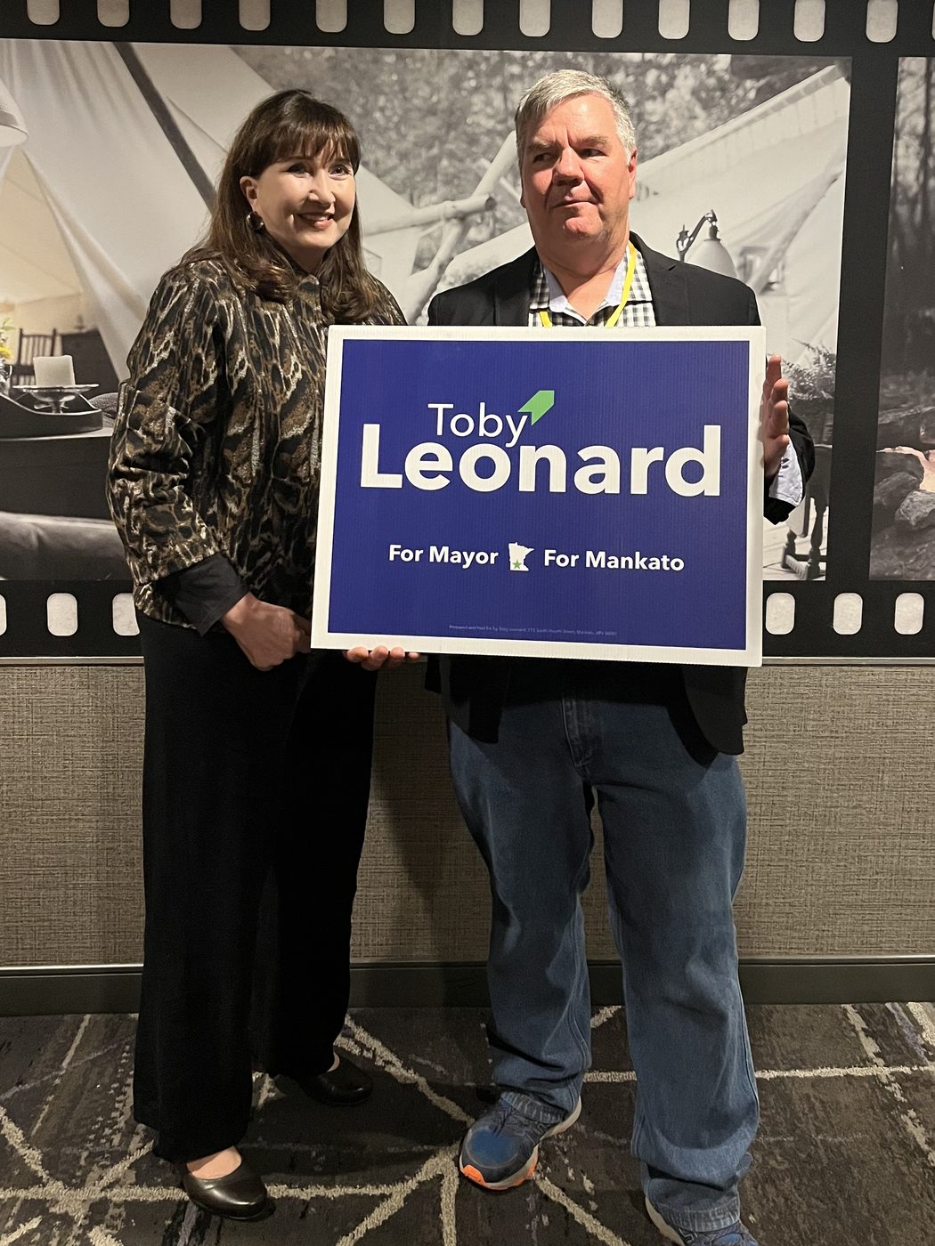 Toby Leonard and a supporter holding a blue 'Toby Leonard For Mayor, For Mankato' yard sign at an indoor event